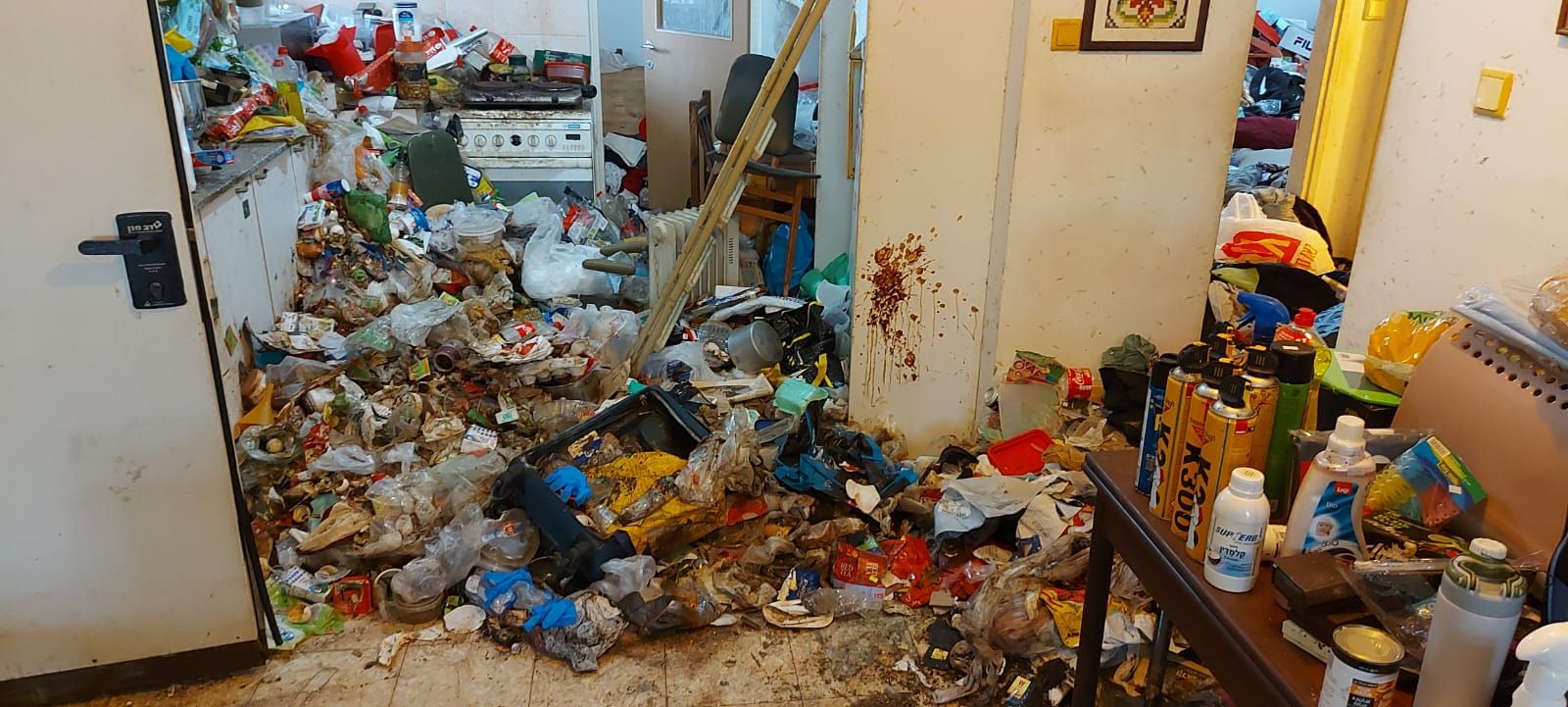 רצפת מטבח מכוסה בפסולת, בקבוקים ושקיות, ועל השולחן בחזית תרסיסים ובקבוקים
