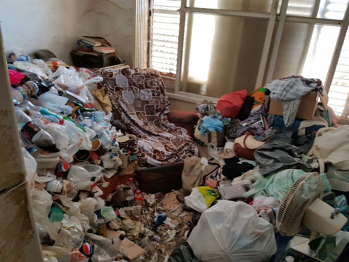 חדר עמוס מקיר לקיר בבקבוקי פלסטיק, בגדים, שמיכות, שקיות ופסולת, ליד חלון עם תריס