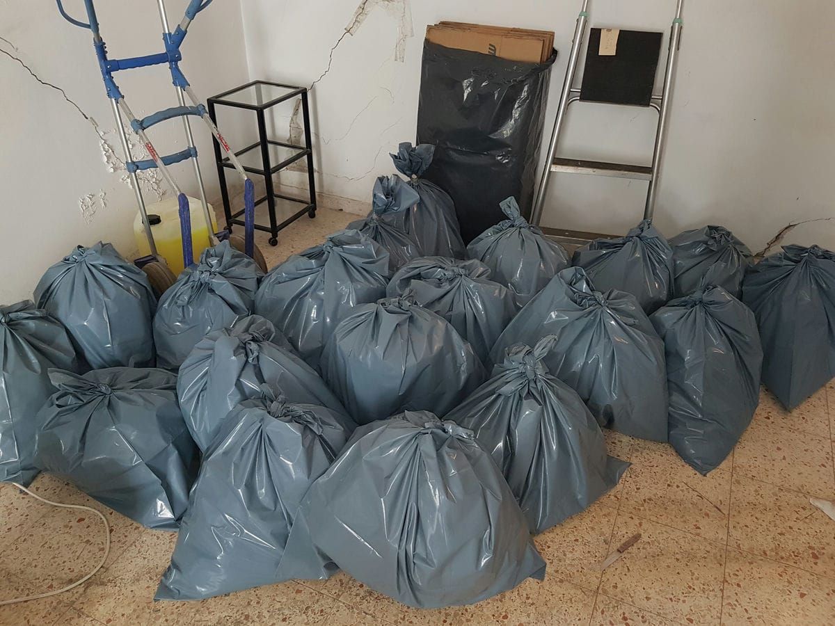 שורות של שקיות תכלת קשורות על רצפת חדר, לצד עגלת משא, מדף מתכת וסולם