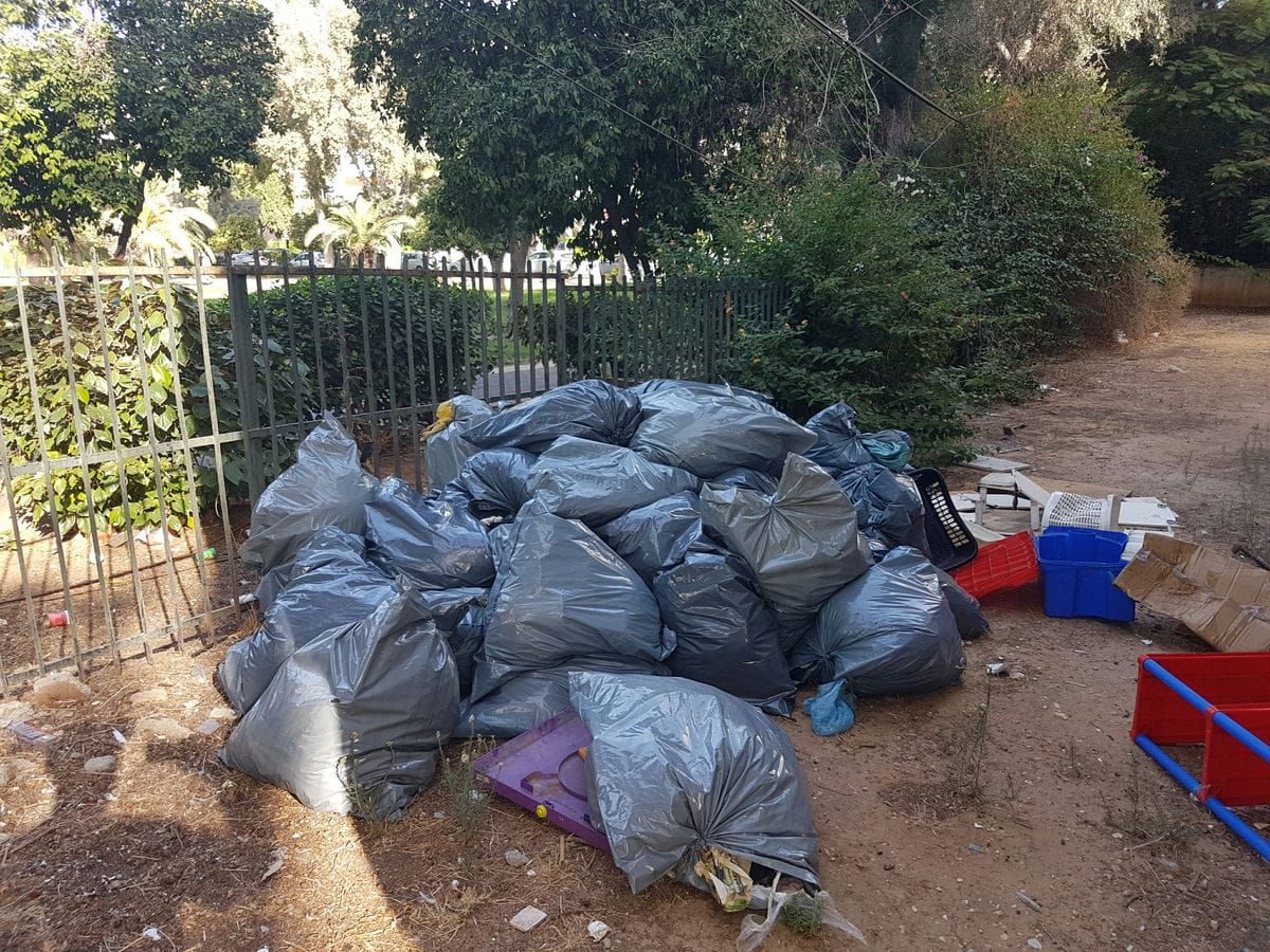 ערימת שקיות אשפה אפורות וארגזי פלסטיק בחצר ליד גדר ועצים