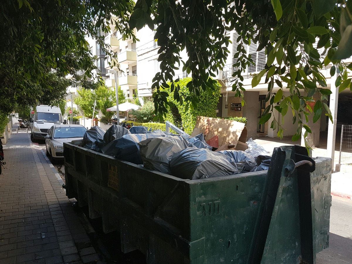 מכולה ירוקה מלאה בשקיות אשפה שחורות על מדרכה ברחוב מוצל