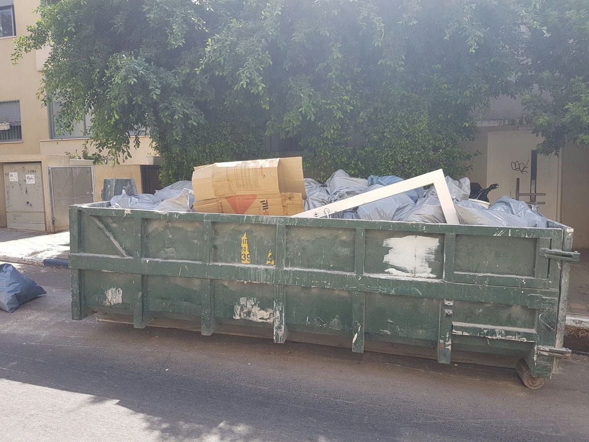 מכולת פסולת ירוקה מלאה בשקיות וקרטונים, עומדת בצד הכביש מול בניין