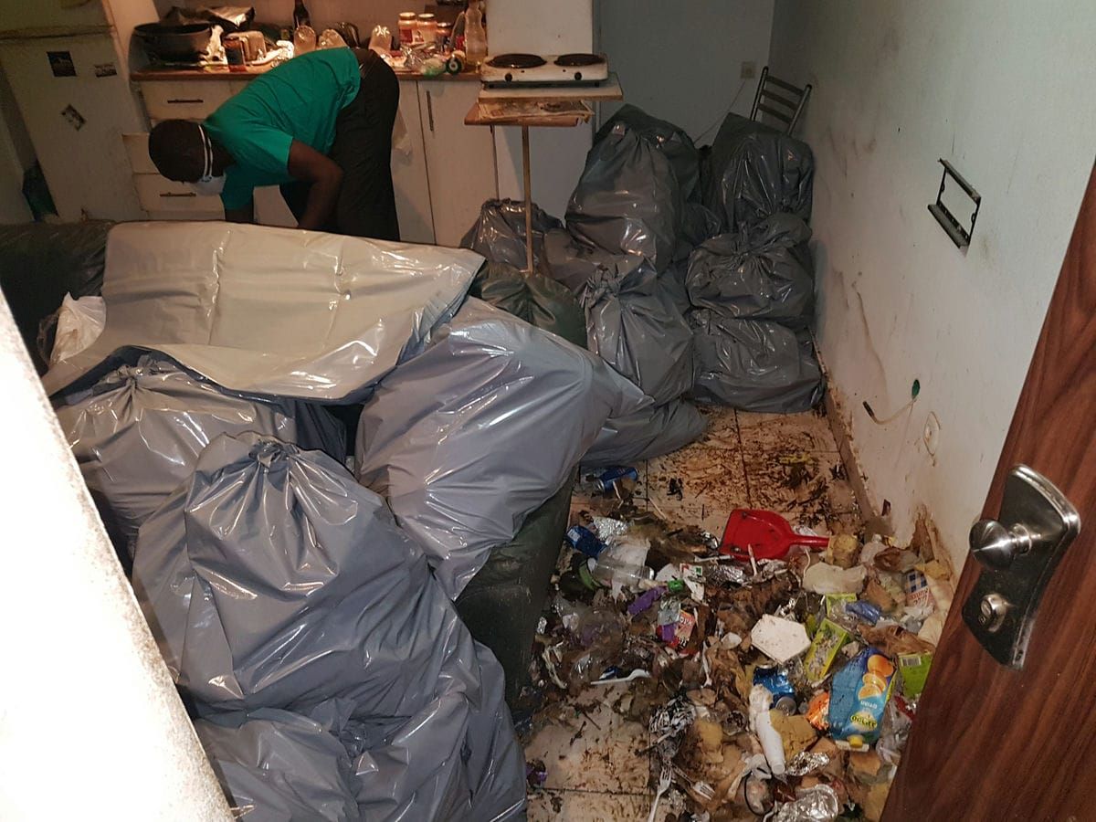 שקיות אשפה מלאות ליד ספה ורצפה מכוסה בפסולת, ועובד עם מסכה ליד המטבחון
