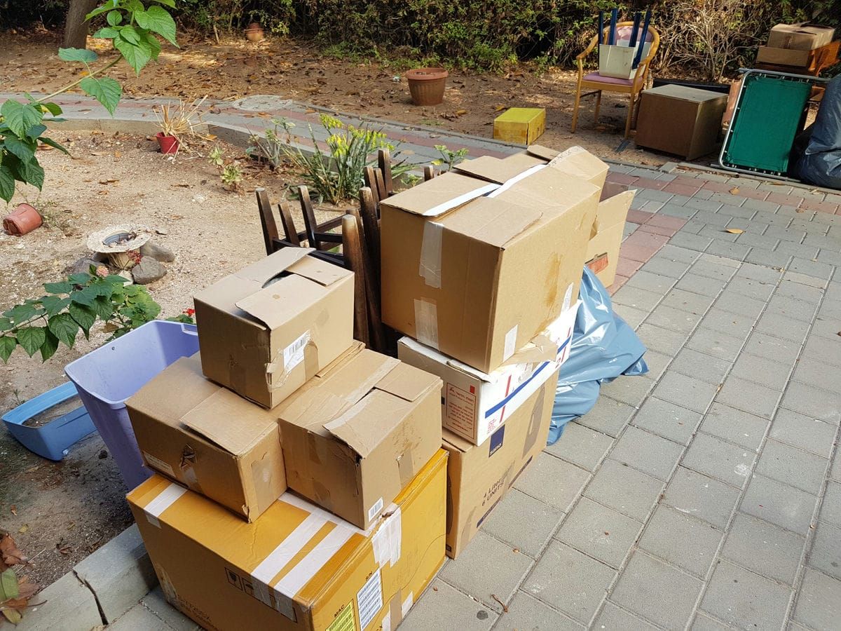 ארגזי קרטון ארוזים, כיסאות וחפצים ערומים על שביל מרוצף ליד גינה