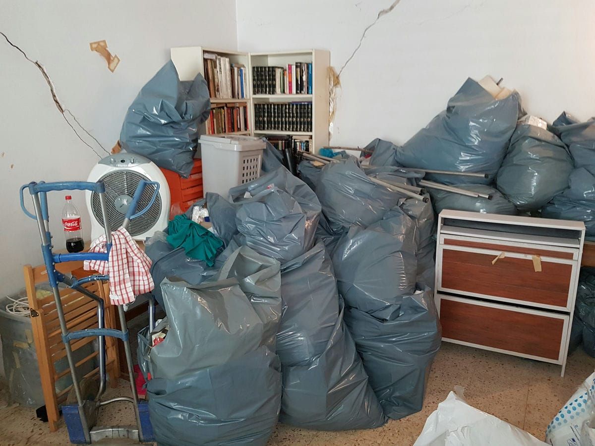 פינת חדר עם שקיות פסולת אפורות מלאות, ספרייה, מאוורר ועגלת משא