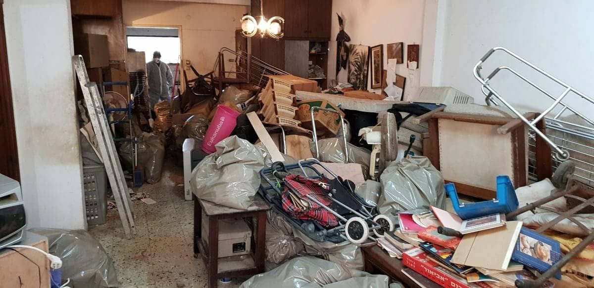 דירה עמוסה בחפצים ושקיות אשפה במהלך הפינוי, עם איש צוות בחליפת מגן ברקע
