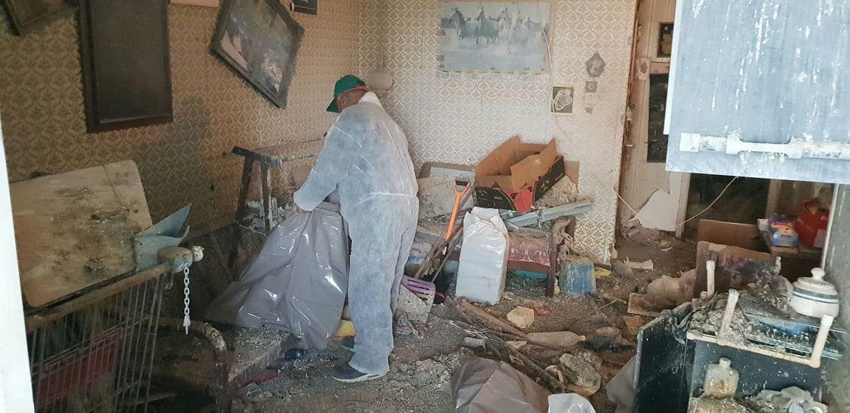 עובד בחליפת מגן ממלא שק גדול בפסולת בחדר עם טפט מתקלף ולכלוך רב על הרצפה