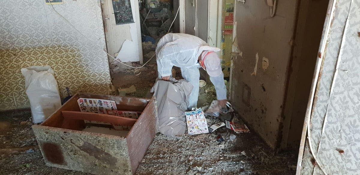 עובד בחליפת מגן אוסף פסולת לשק ליד מגירה ישנה, בחדר שרצפתו מכוסה פסולת