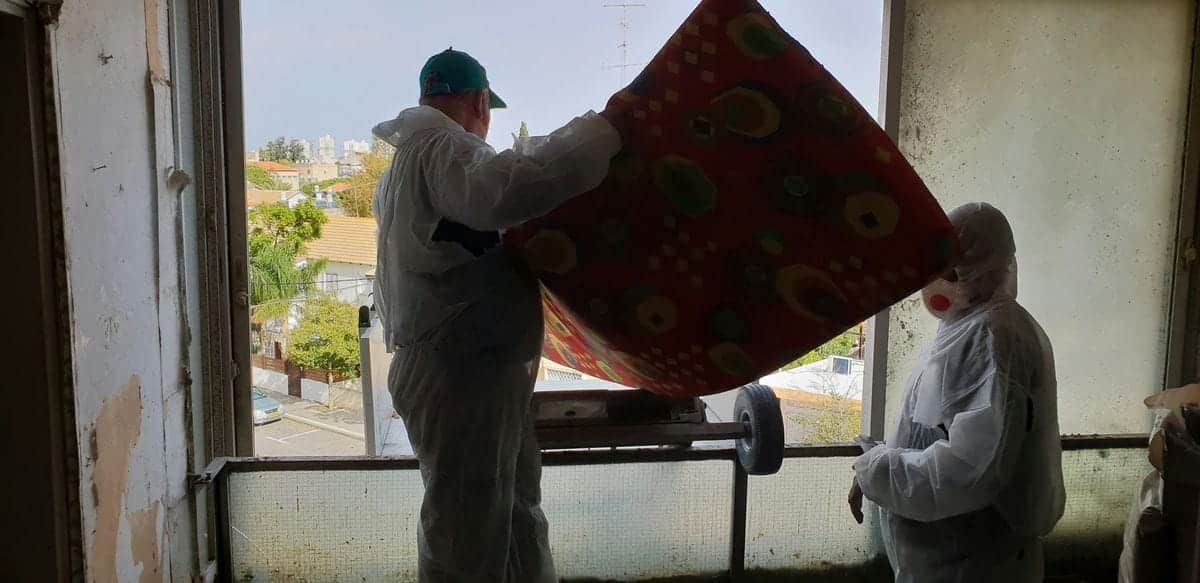 שני עובדים בחליפות מגן מעמיסים מזרן על מנוף הרמה דרך פתח חלון במרפסת